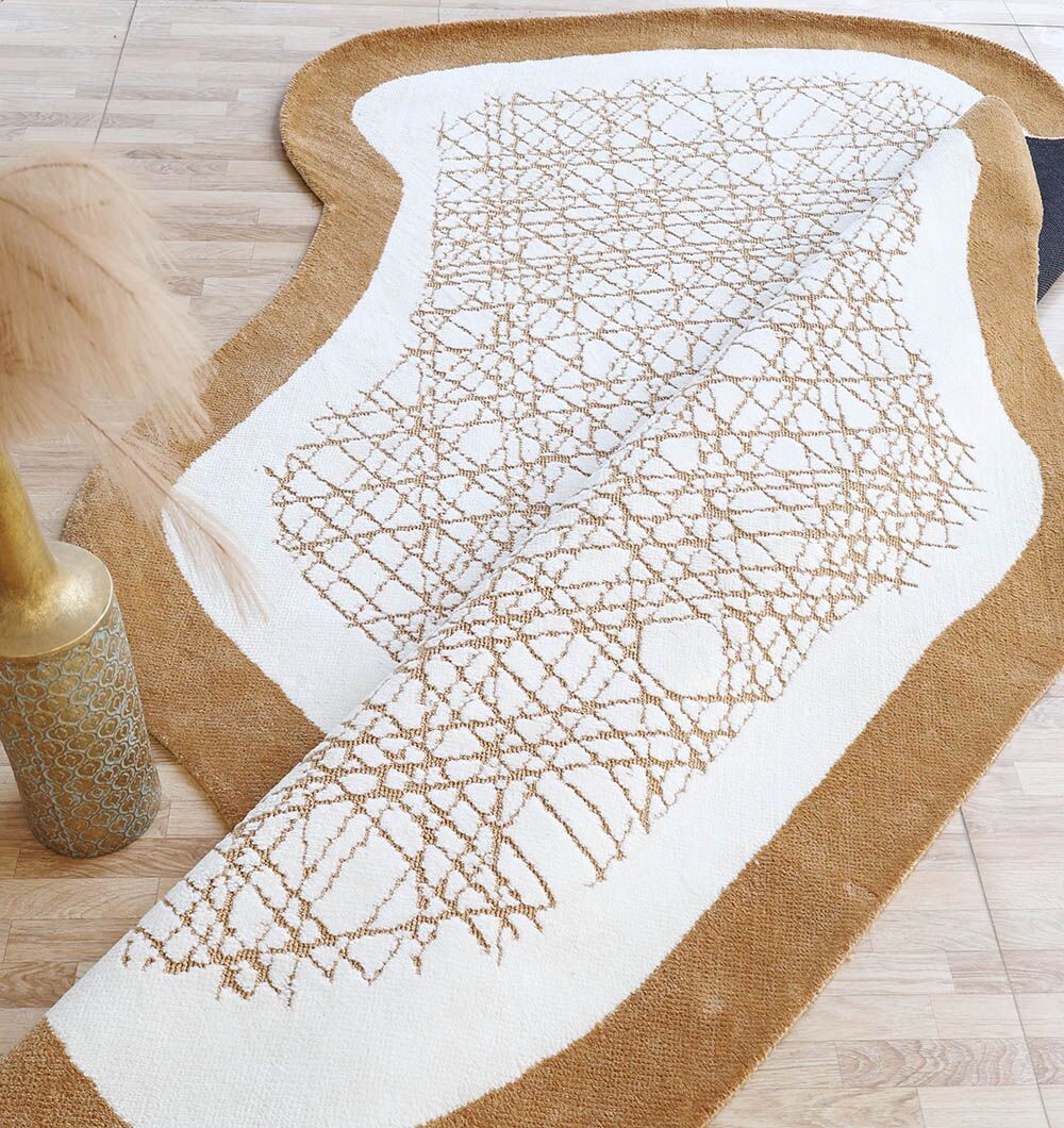 The Novara Shape White & Beige Area Rug (200x300 cm) by RAMSHA HOME features a white center, tan border, and brown crisscross design. Partially folded on a light wood floor, it's paired with a gold-patterned vase of dried pampas grass for modern appeal.