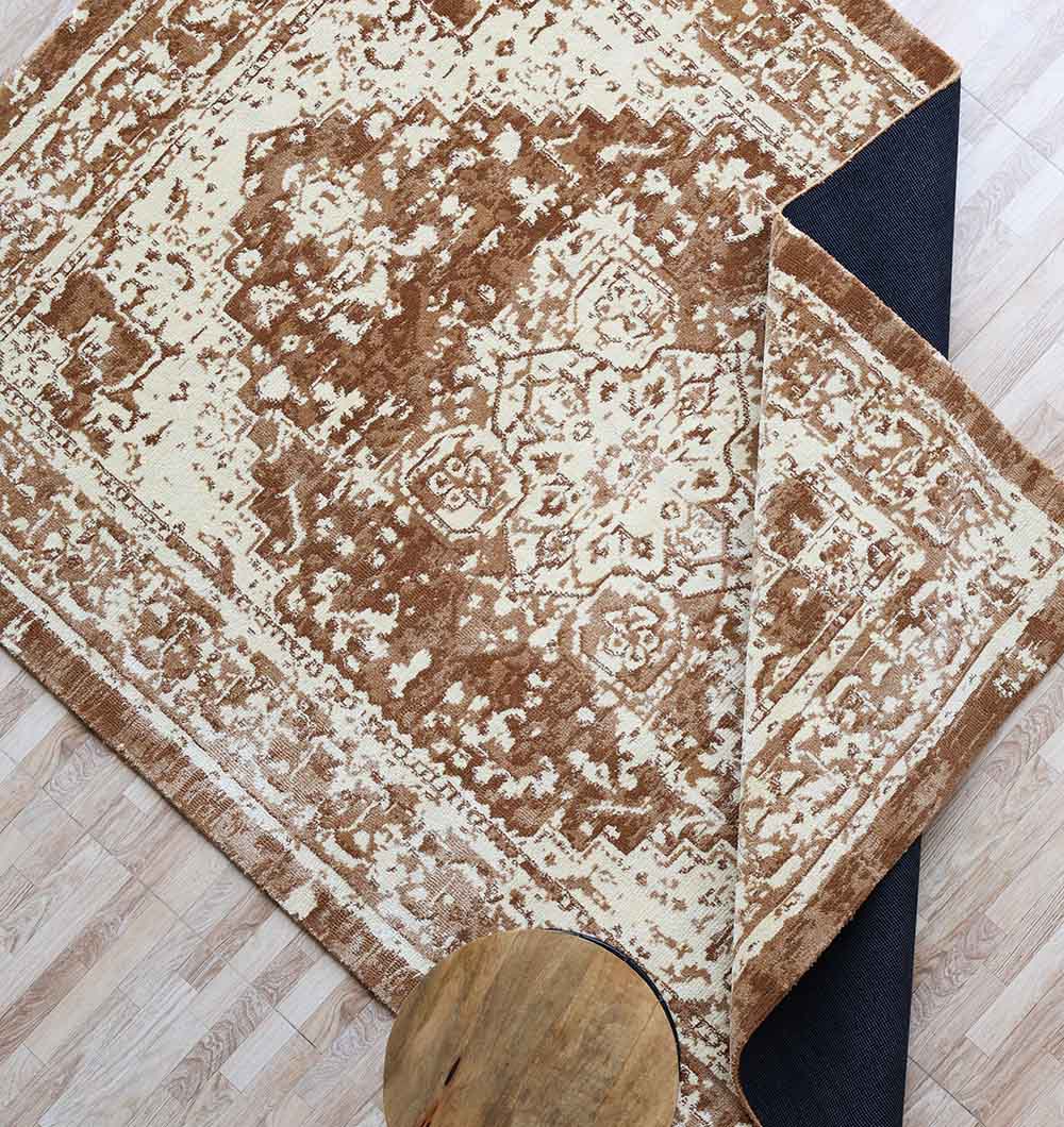 The RAMSHA HOME Traditional Brown Medallion Novara Carpet - CMN 91 (170x240cm) with a black non-slip backing is displayed on a light wood floor, its corner flipped over. A small round wooden table sits nearby.