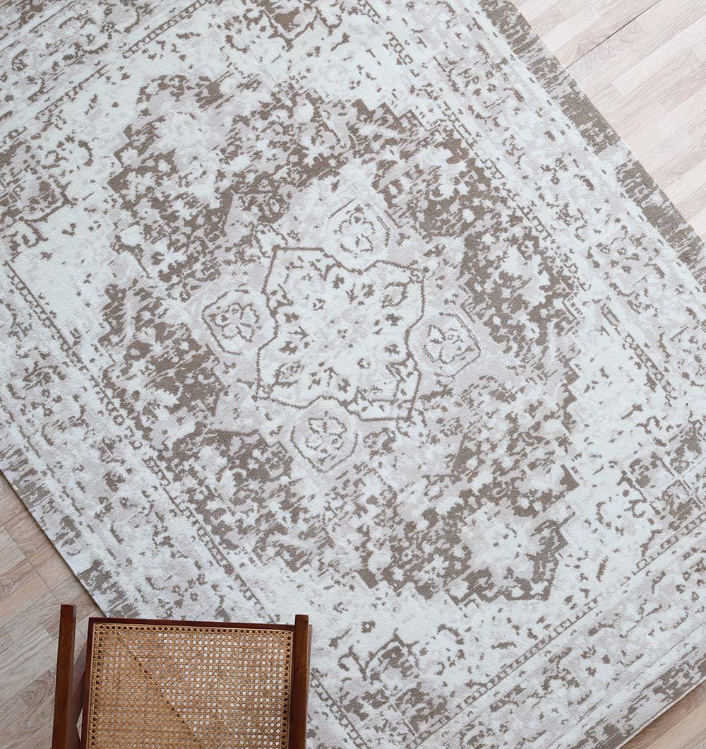 The RAMSHA HOME Grey Vintage Style Novara Carpet - CMN 92 features a faded, ornate central medallion on a wood floor with a wooden chair and woven rattan seat in the lower left corner.