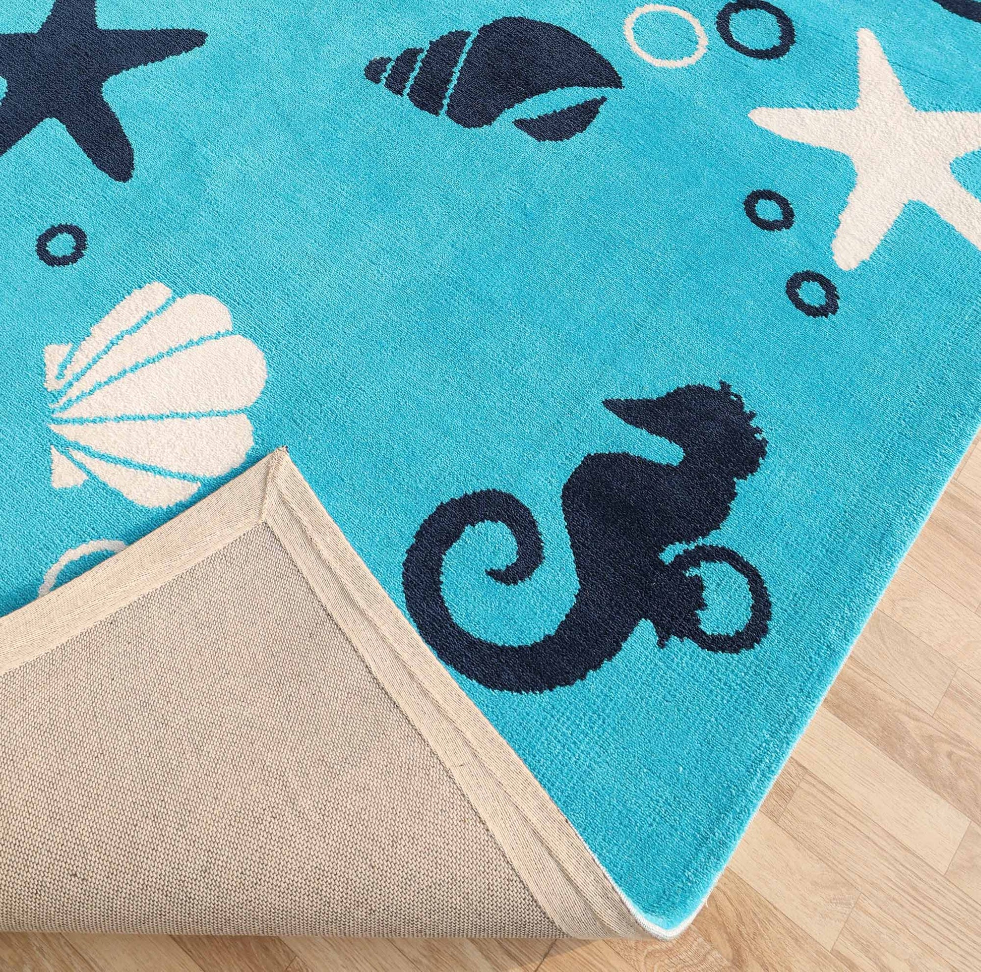 The RAMSHA HOME Ultra Soft Ocean Animals Kids Rug (CMN 100, 200x300 cm) features white and navy sea creatures on a blue background. Its corner is flipped to show the beige underside, adding playful charm to any kids room with a wooden floor.