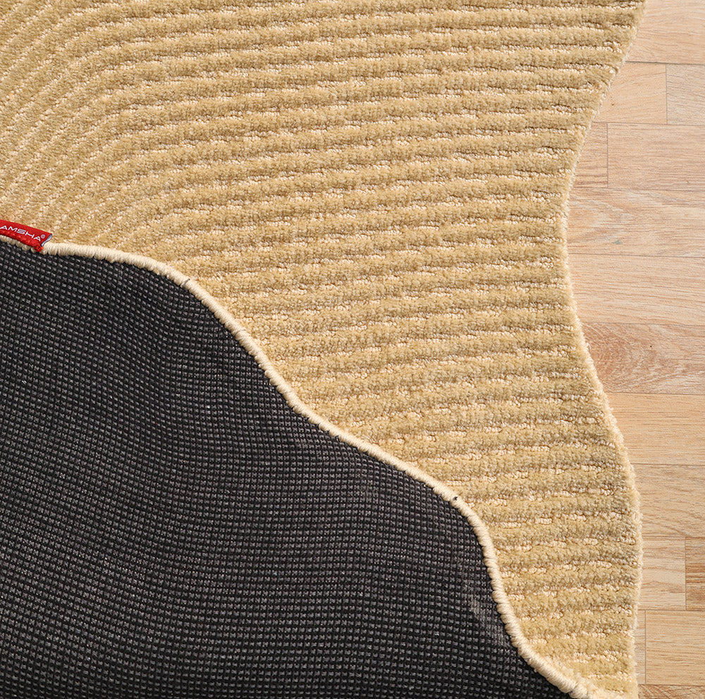 A close-up of the RAMSHA HOME Dune Flow Abstract Shaped Rug Beige & Terracotta - CMN 102 shows its wavy edges on a wooden floor, partially lifted to reveal the dark non-slip backing and a small red fabric tag.