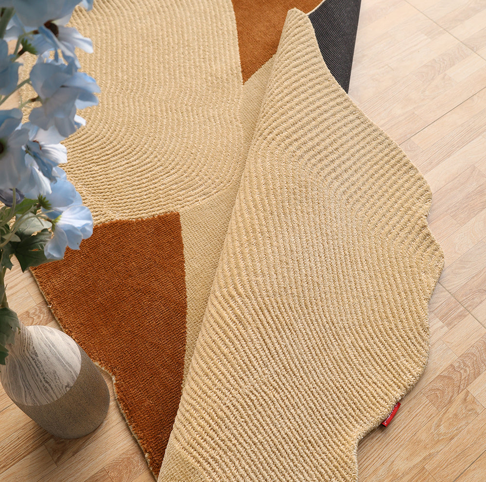 The RAMSHA HOME Dune Flow Abstract Shaped Rug in beige and terracotta (CMN 102) features a modern design and is partly folded on a light wood floor, with a vase of white flowers visible on the left.