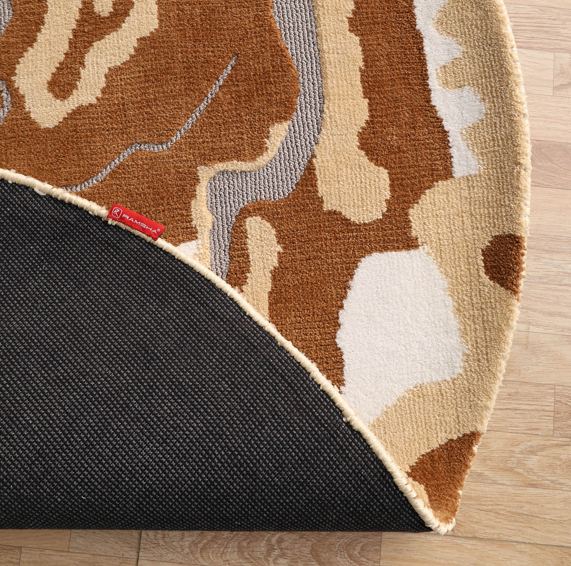 A close-up of the RAMSHA HOME Modern Round Abstract Rug CMN 104 shows tan, cream, and brown shapes. The rug’s flipped corner reveals a black textured underside with a red tag, resting on a light wood floor.