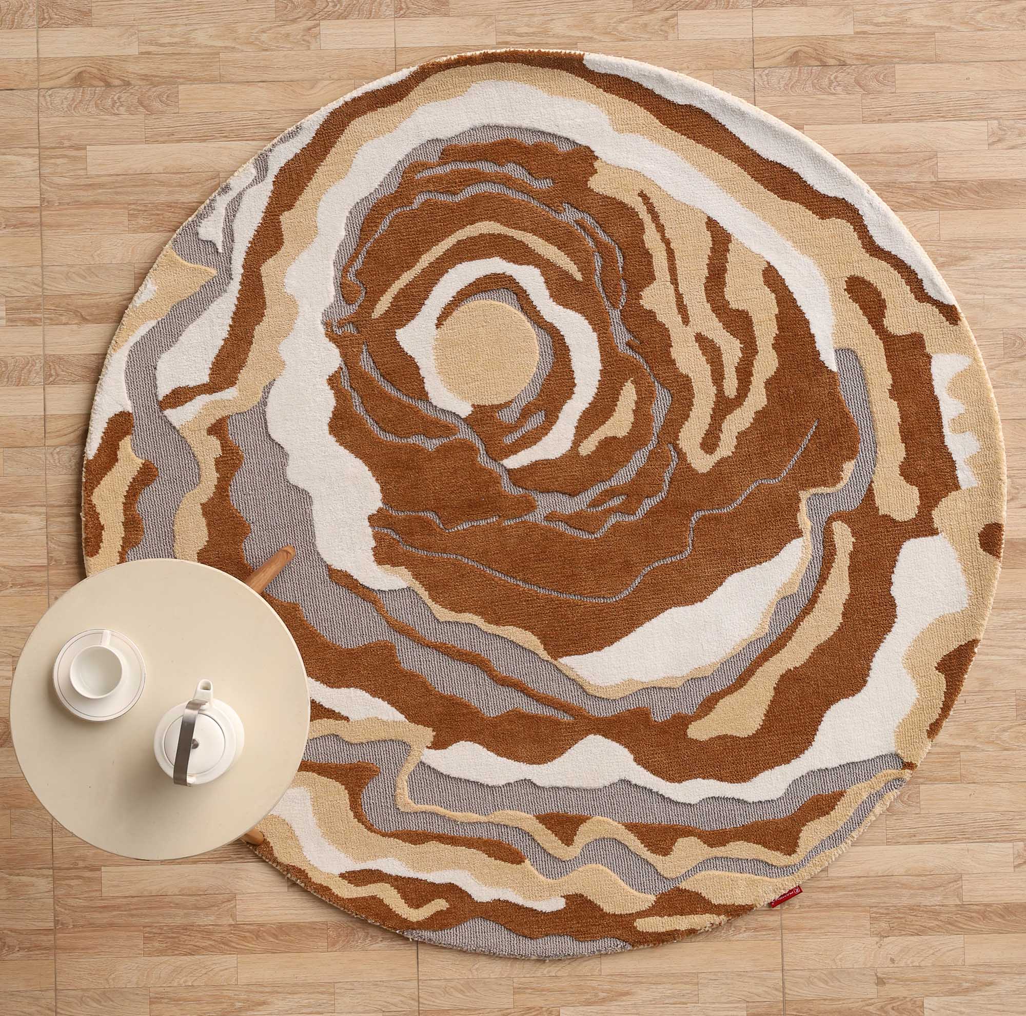 The RAMSHA HOME Modern Round Abstract Rug CMN 104, featuring swirling brown, beige, white, and gray patterns, lies on a light wood floor with a small beige table holding two white teacups and saucers partly atop the rug.
