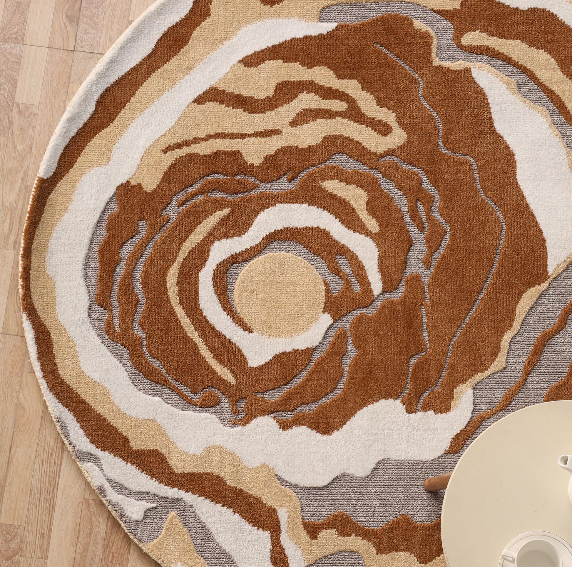 The Modern Round Abstract Rug CMN 104 by RAMSHA HOME, featuring swirling brown, beige, and gray patterns, is displayed on a wooden floor beside a light table and a visible hand at the image’s edge.