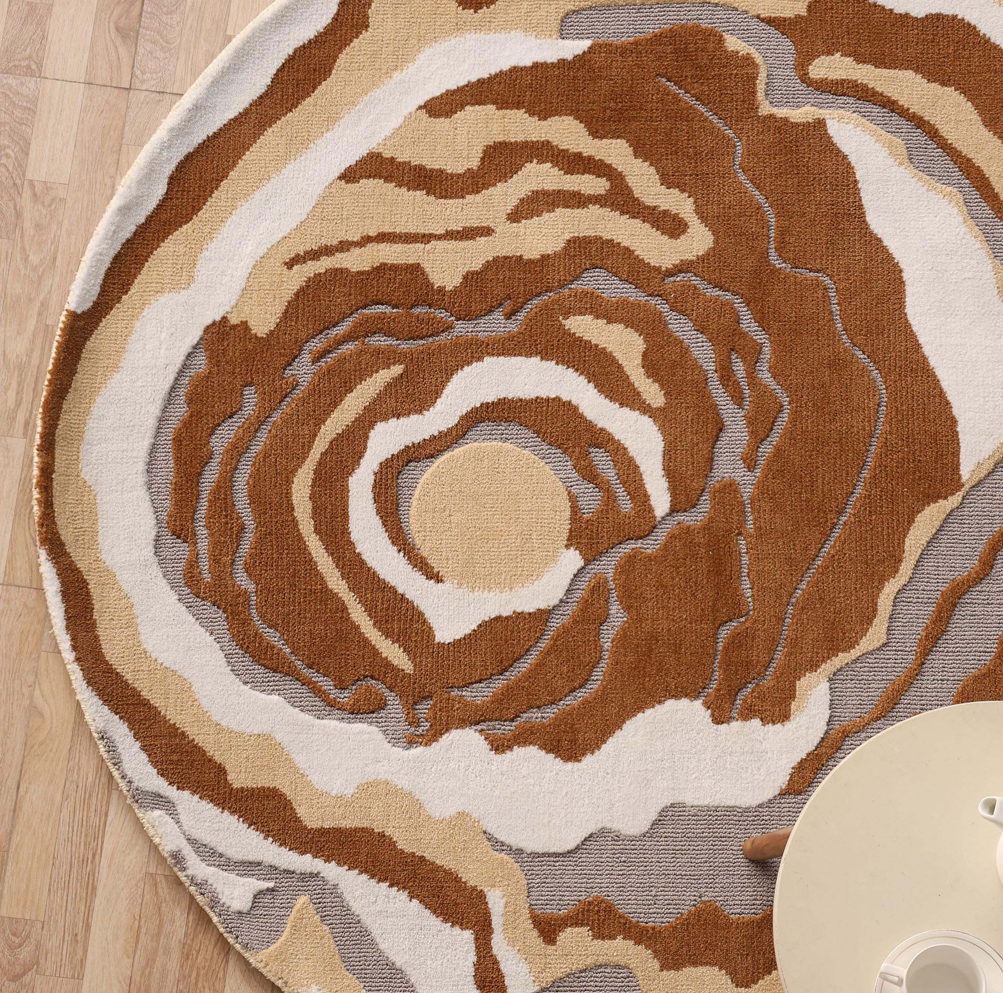 The Modern Round Abstract Rug CMN 104 by RAMSHA HOME, featuring swirling brown, beige, and gray patterns, is displayed on a wooden floor beside a light table and a visible hand at the image’s edge.