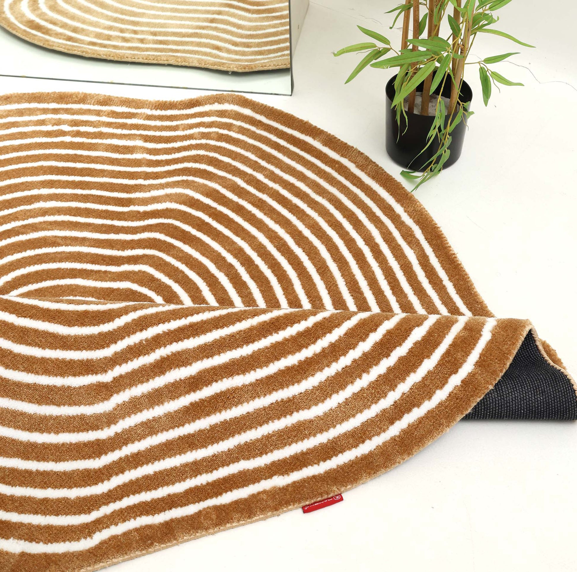The RAMSHA HOME Contour Flow Abstract Shaped Rug – Camel & Ivory CMN 105 features contemporary curved lines, is partially flipped at one corner, and sits on a white floor beside a potted plant and a mirror reflecting its premium materials.