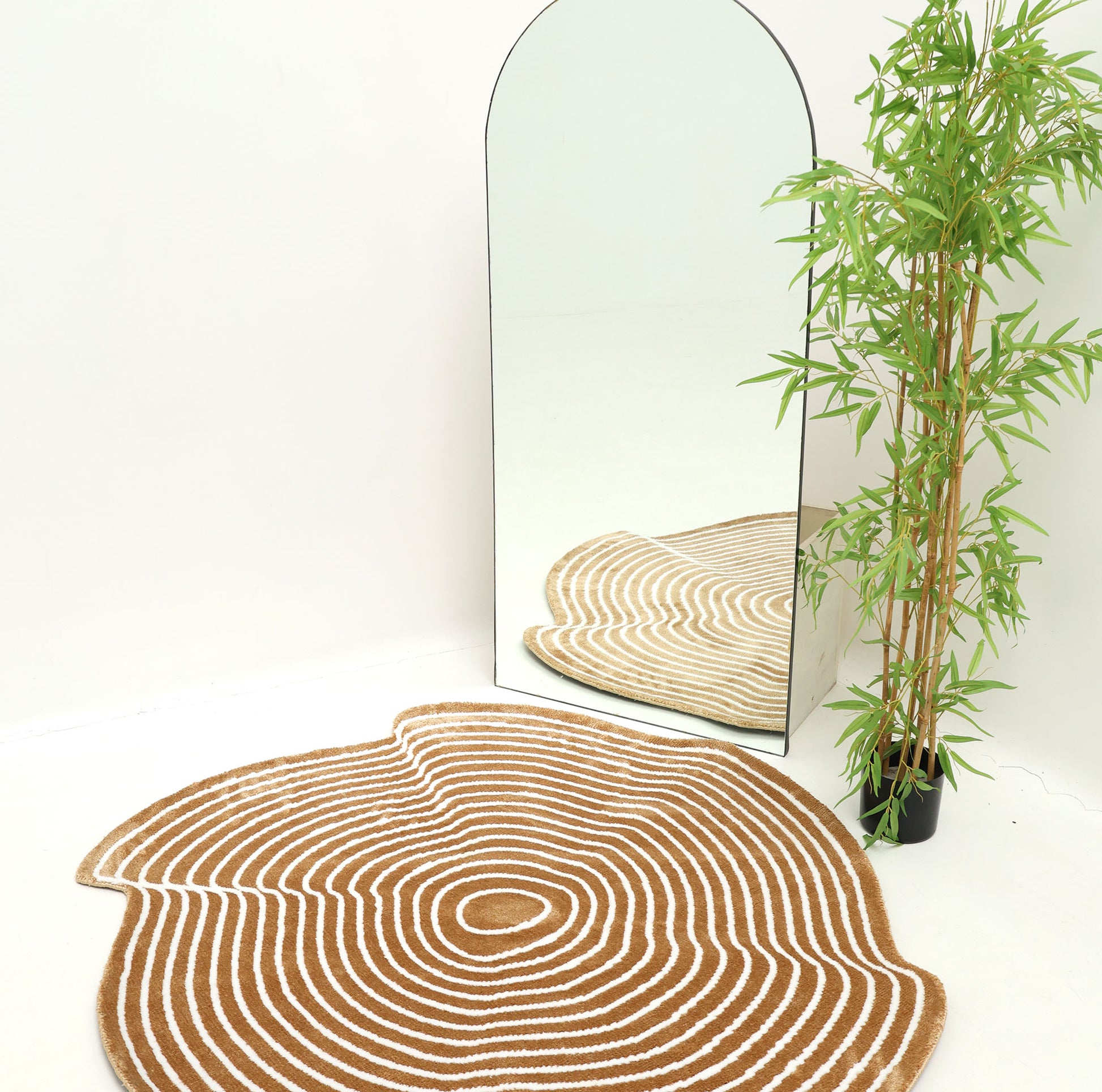 The RAMSHA HOME Contour Flow Abstract Shaped Rug – Camel & Ivory CMN 105, featuring tree ring-like concentric rings, lies on a white floor beside an arched mirror and potted bamboo, with the mirror reflecting the rug and minimalist decor.