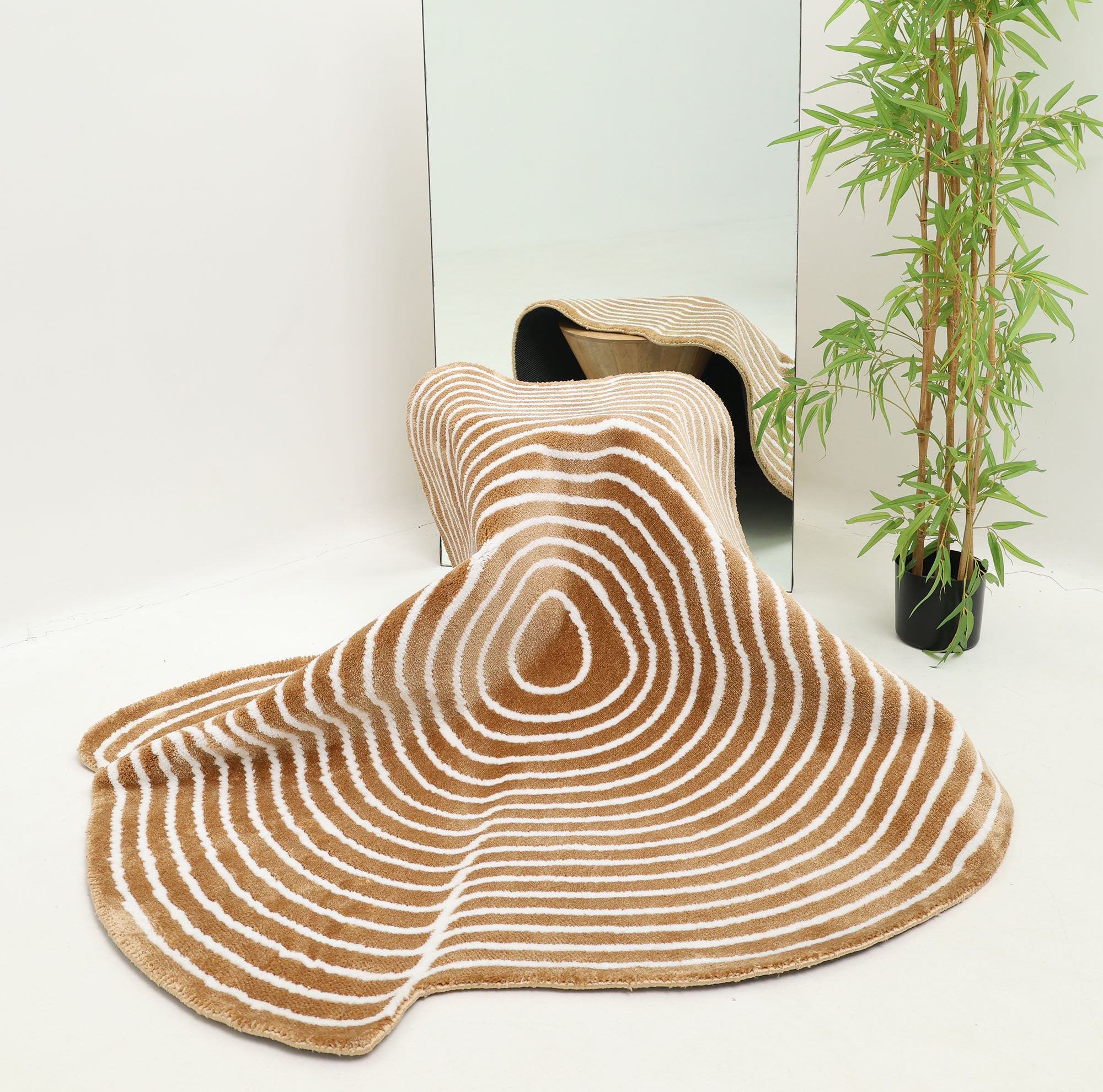 The RAMSHA HOME Contour Flow Abstract Shaped Rug – Camel & Ivory CMN 105, with tan and white stripes, is draped in front of a mirror for a wavy effect. A tall green plant sits nearby in this minimal space with white walls and floor.