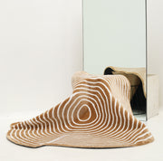 The Contour Flow Abstract Shaped Rug in Camel & Ivory by RAMSHA HOME, with stripe and oval patterns, drapes over a wire basket before a large mirror, creating a distorted reflection in this minimal, white-walled room.