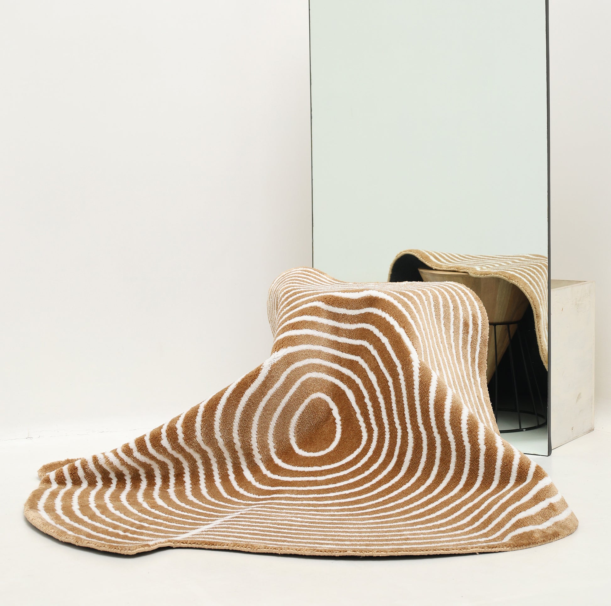 The Contour Flow Abstract Shaped Rug in Camel & Ivory by RAMSHA HOME, with stripe and oval patterns, drapes over a wire basket before a large mirror, creating a distorted reflection in this minimal, white-walled room.