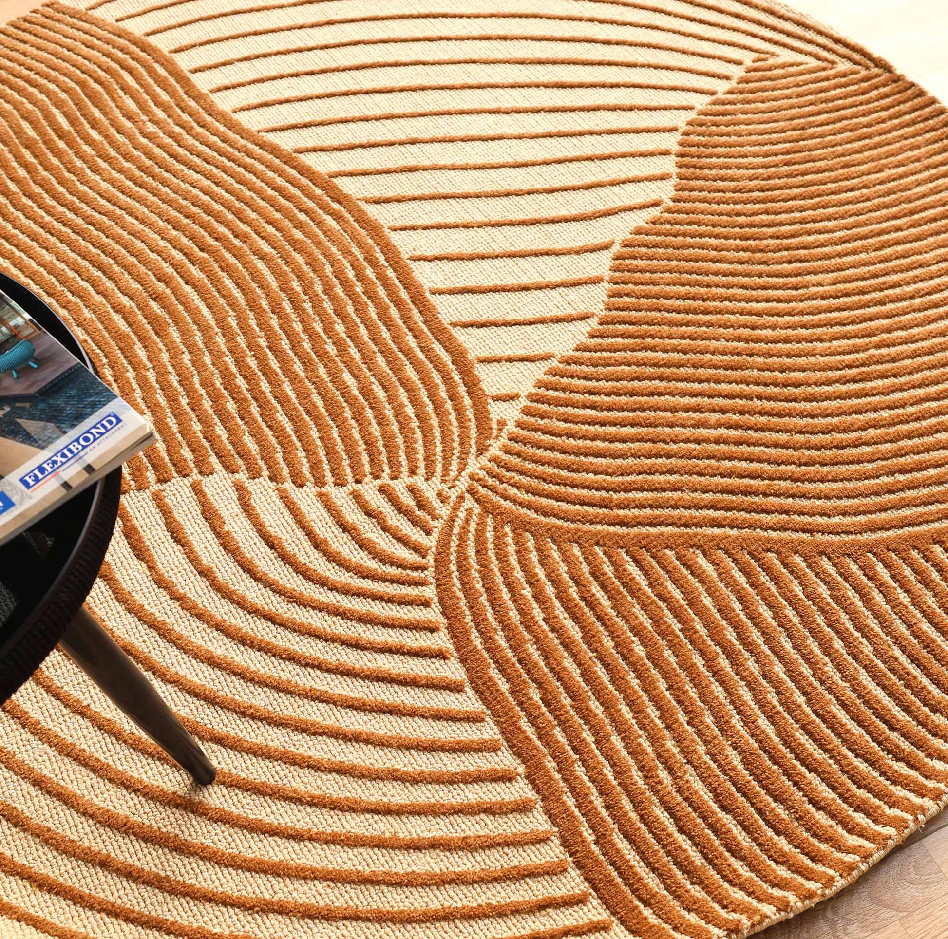The Layered Wave Modern Round Abstract Rug CMN 106 by RAMSHA HOME, with textured beige and brown wavy patterns, is shown beside a black round table with a partially visible book—ideal for lovers of contemporary design.