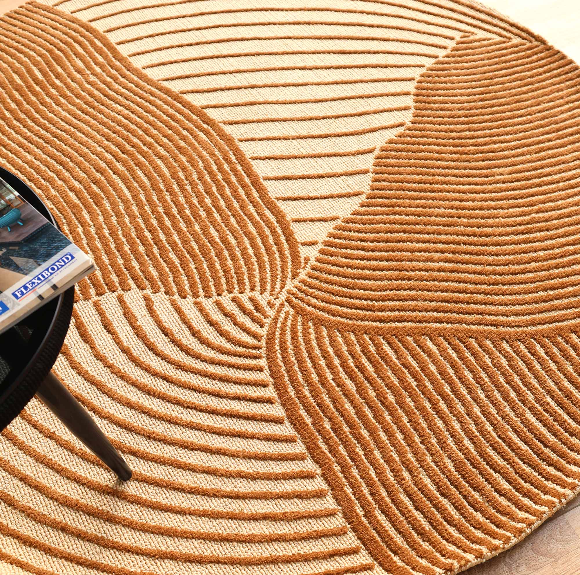 The Layered Wave Modern Round Abstract Rug CMN 106 by RAMSHA HOME, with textured beige and brown wavy patterns, is shown beside a black round table with a partially visible book—ideal for lovers of contemporary design.