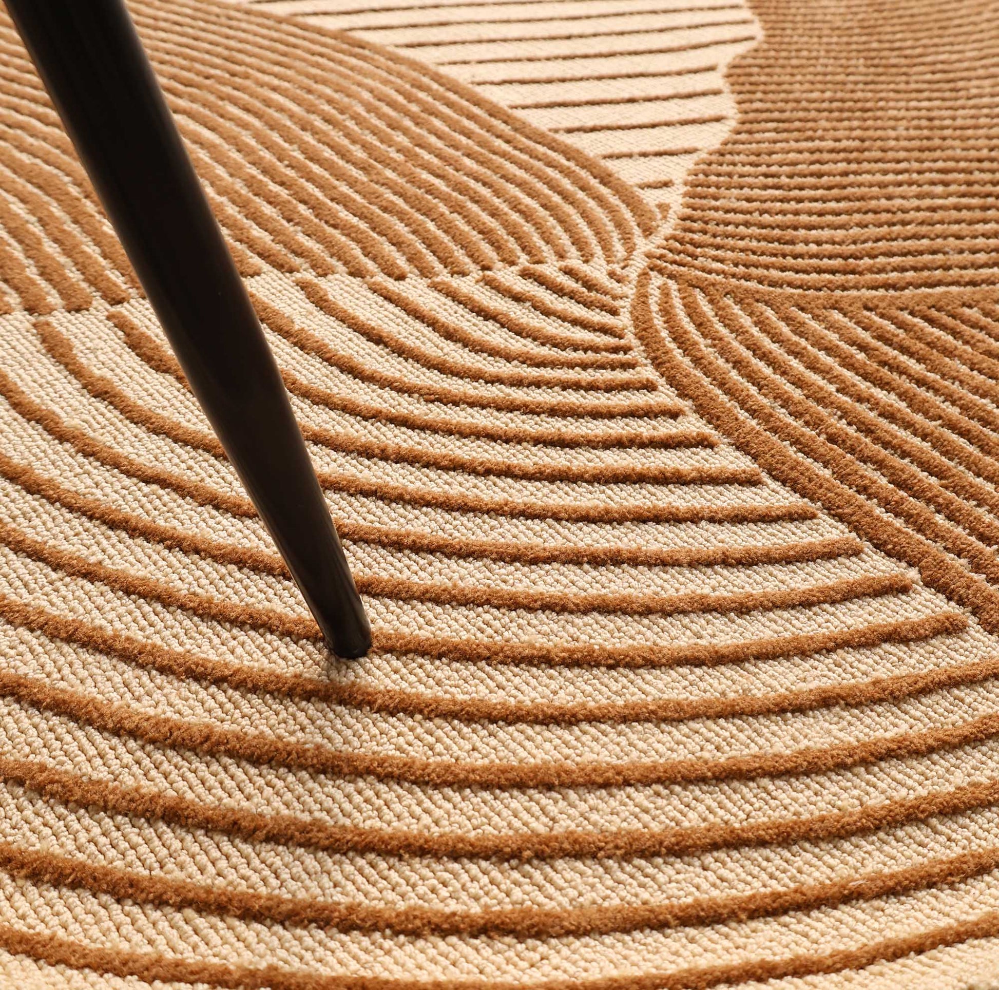 A close-up of the RAMSHA HOME Layered Wave Modern Round Abstract Rug CMN 106 in beige and brown, featuring wavy raised patterns, geometric accents, and intersected by a slim black furniture leg.