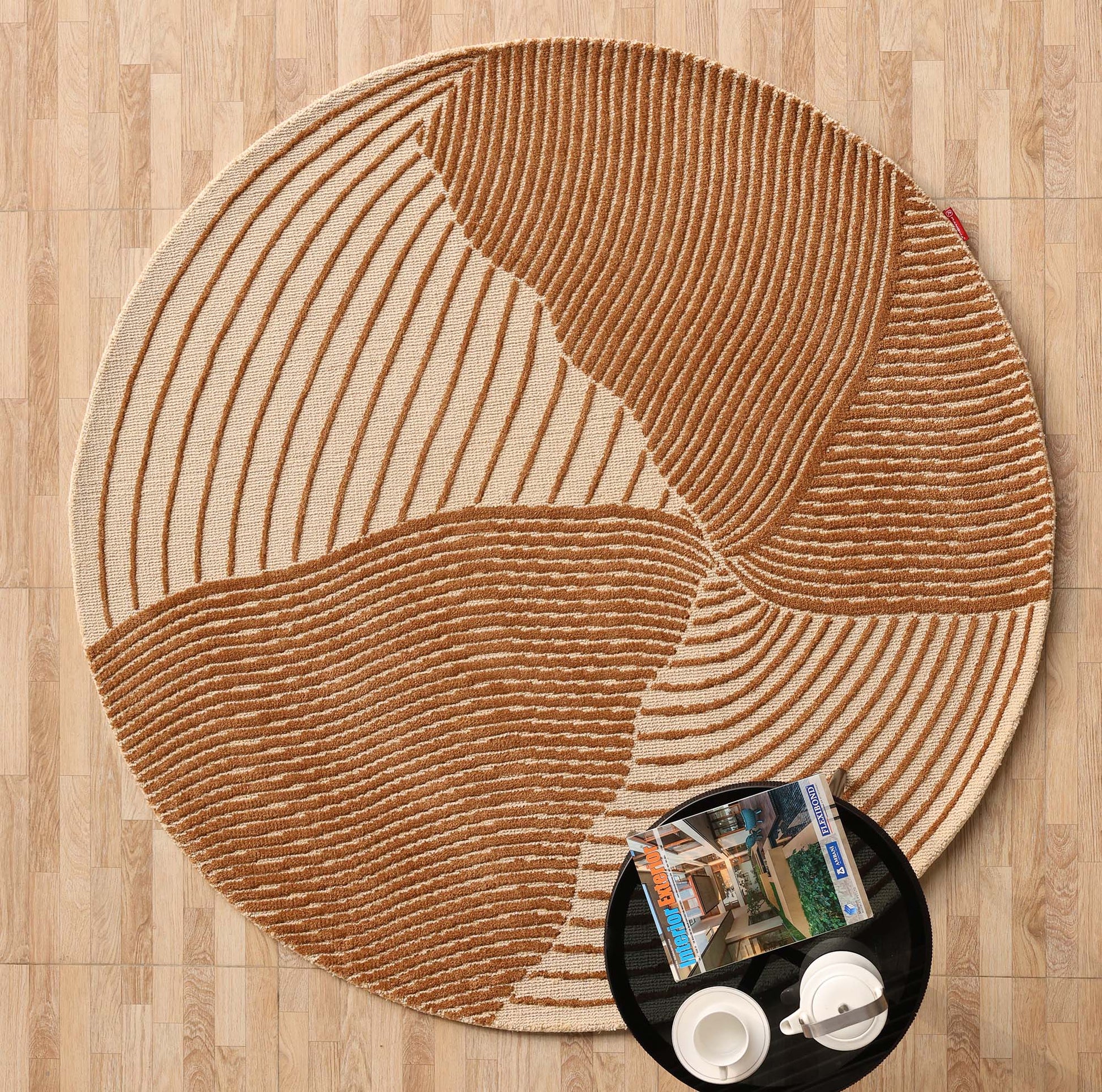 The Layered Wave Modern Round Abstract Rug CMN 106 by RAMSHA HOME features beige and brown textured wavy lines on wood flooring, with a black tray holding white cups and magazines, enhancing the contemporary look.
