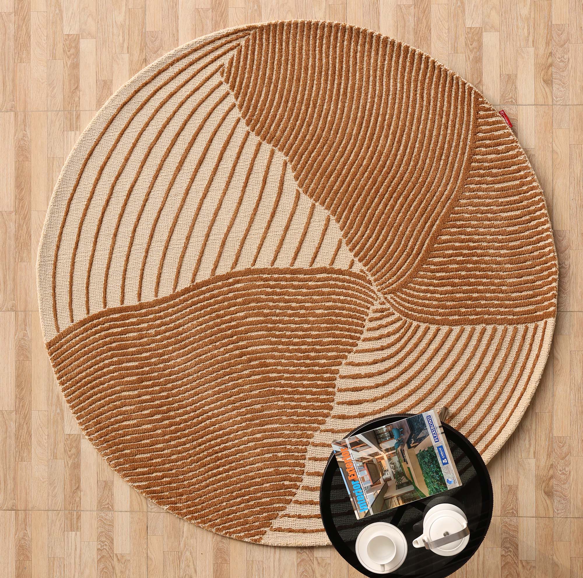 The Layered Wave Modern Round Abstract Rug CMN 106 by RAMSHA HOME features beige and brown textured wavy lines on wood flooring, with a black tray holding white cups and magazines, enhancing the contemporary look.