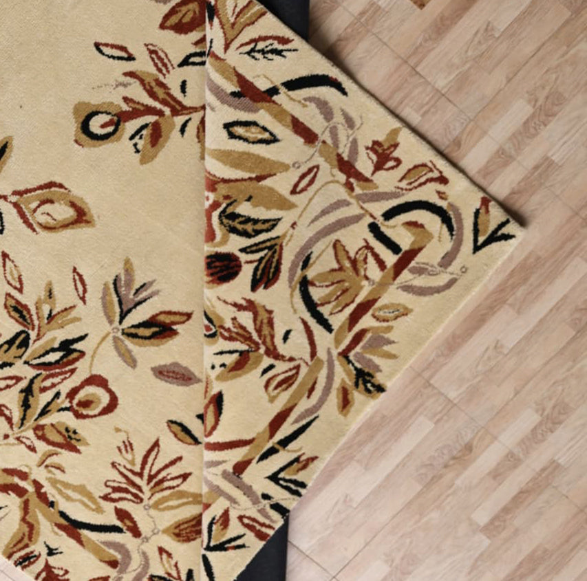 The RAMSHA HOME Vintage Floral Elegance Handmade Rug 200x300 CM – CMN 111 in beige is partially folded at the corner, revealing a black rug pad beneath it on a light wood floor.