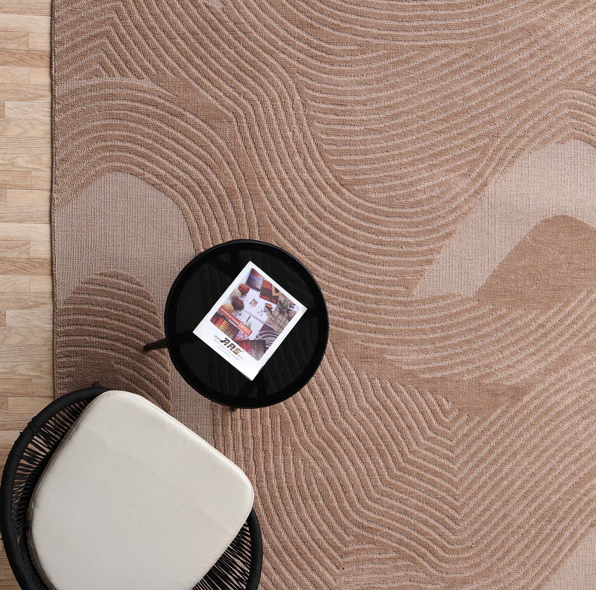 The RAMSHA HOME Terrain Design 200x300 CM Rectangle Shaped Rug CMN 112 in beige with wavy patterns is placed on a wooden floor, with a black round side table holding a magazine and a chair with a white cushion set atop the rug.