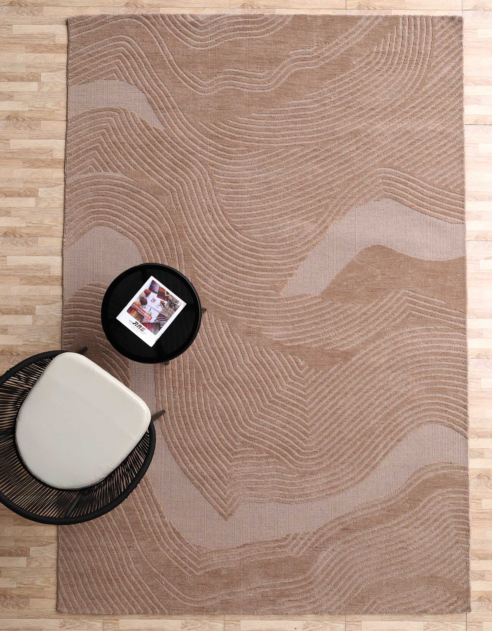 The RAMSHA HOME Terrain Design 200x300 CM Rectangle Shaped Rug CMN 112 in beige and tan with abstract wavy lines lies on a light wood floor, next to a round black side table and a woven black chair with a white cushion.