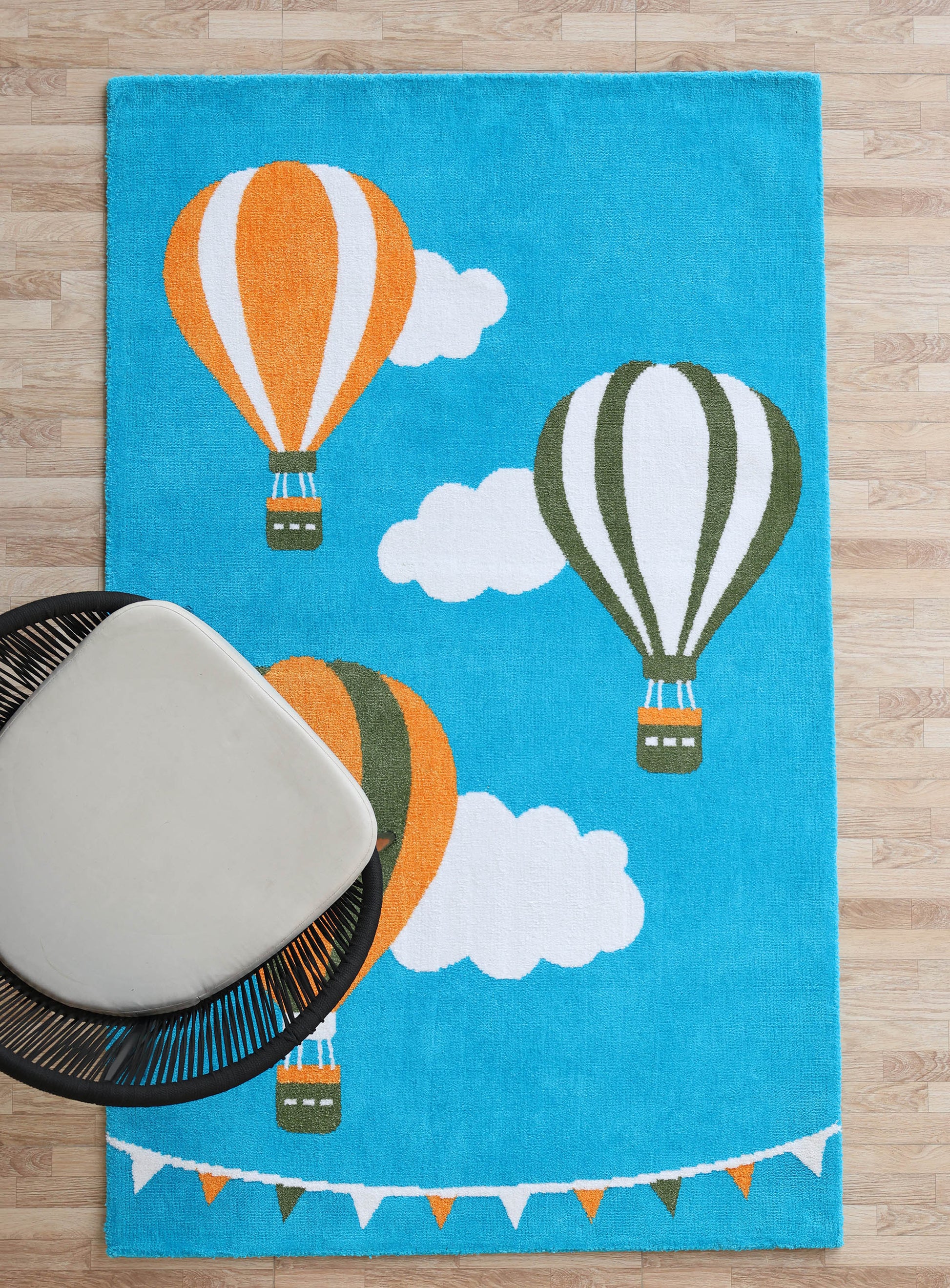 The RAMSHA HOME Kids Play Rug with Clouds & Balloons (CMN 101, 200x300 CM) adds playful balloon, cloud, and pennant flag illustrations to the room. A modern white chair rests partly on this charming rug over a light wood floor.