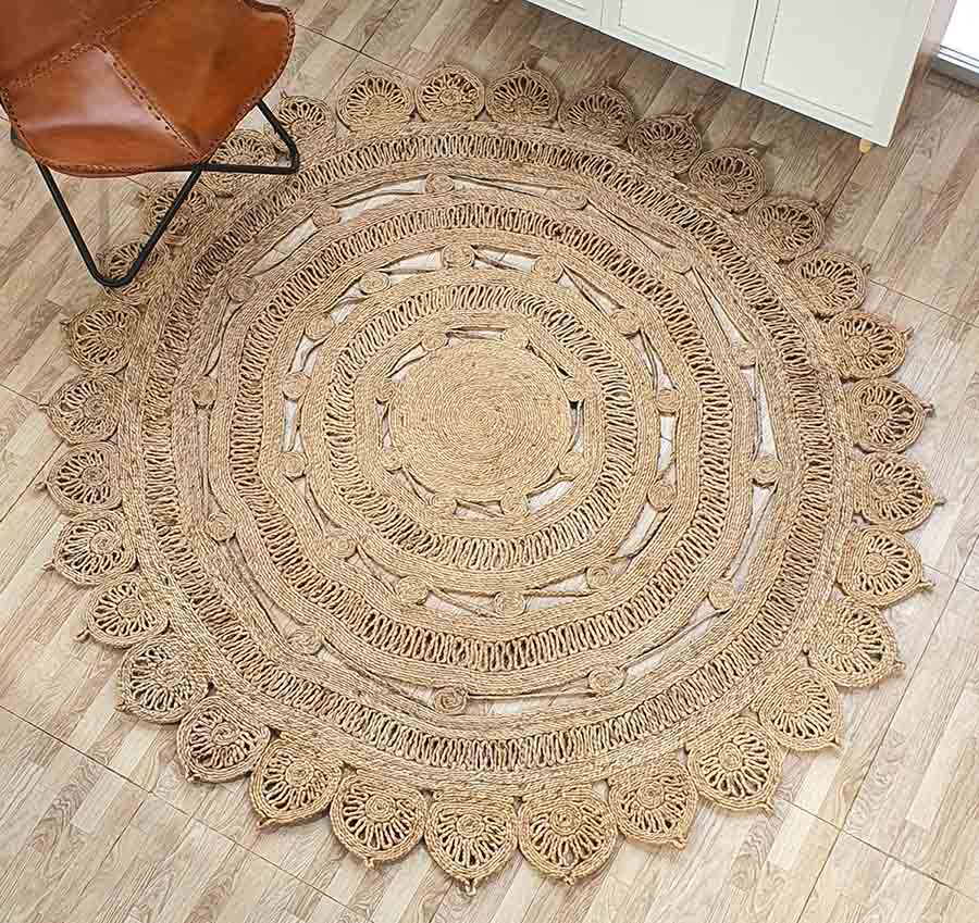The RAMSHA HOME Natural Crochet Jute Round Rug - CR 013, featuring intricate lacy patterns and scalloped edges, is displayed on a light wood floor beside a brown leather chair and a white cabinet.