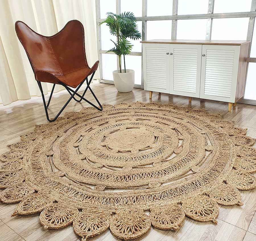 The RAMSHA HOME Natural Crochet Jute Round Rug - CR 013 lies on a wooden floor near a brown leather chair, white louvered cabinet, potted plant, and large windows with sheer curtains.