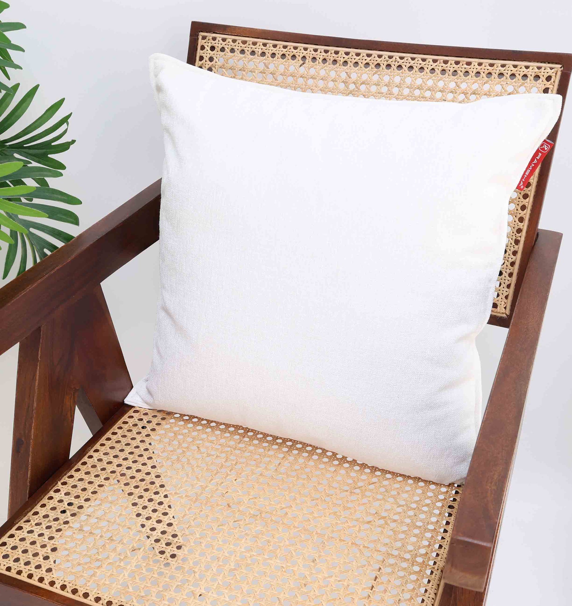 The White Color Decorative Cushion For Sofa - CS 293 by Ramsha Home adds elegance to a wooden chair with a cane seat, while a green plant on the side enhances freshness against a plain white background. Perfect for stylish, modern interiors.
