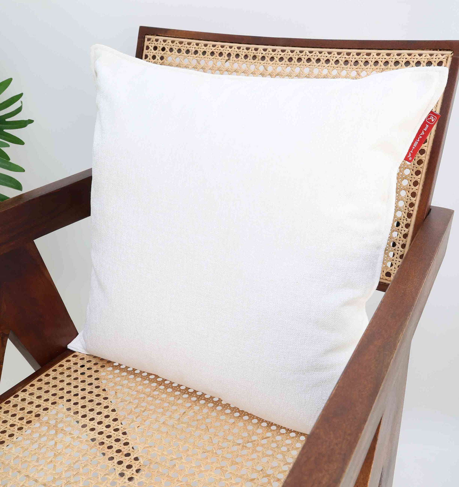 The White Color Decorative Cushion For Sofa - CS 293 by Ramsha Home adds elegance to your space, shown on a wooden chair with cane backrest and seat. A green plant in the background brings fresh style to your home décor.