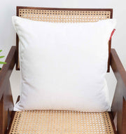 The Ramsha Home White Color Decorative Cushion For Sofa (CS 293) sits on a wooden chair with woven cane, its subtle red tag adding charm to the scene. A green plant peeks in from the left, enhancing this inviting home décor setting.