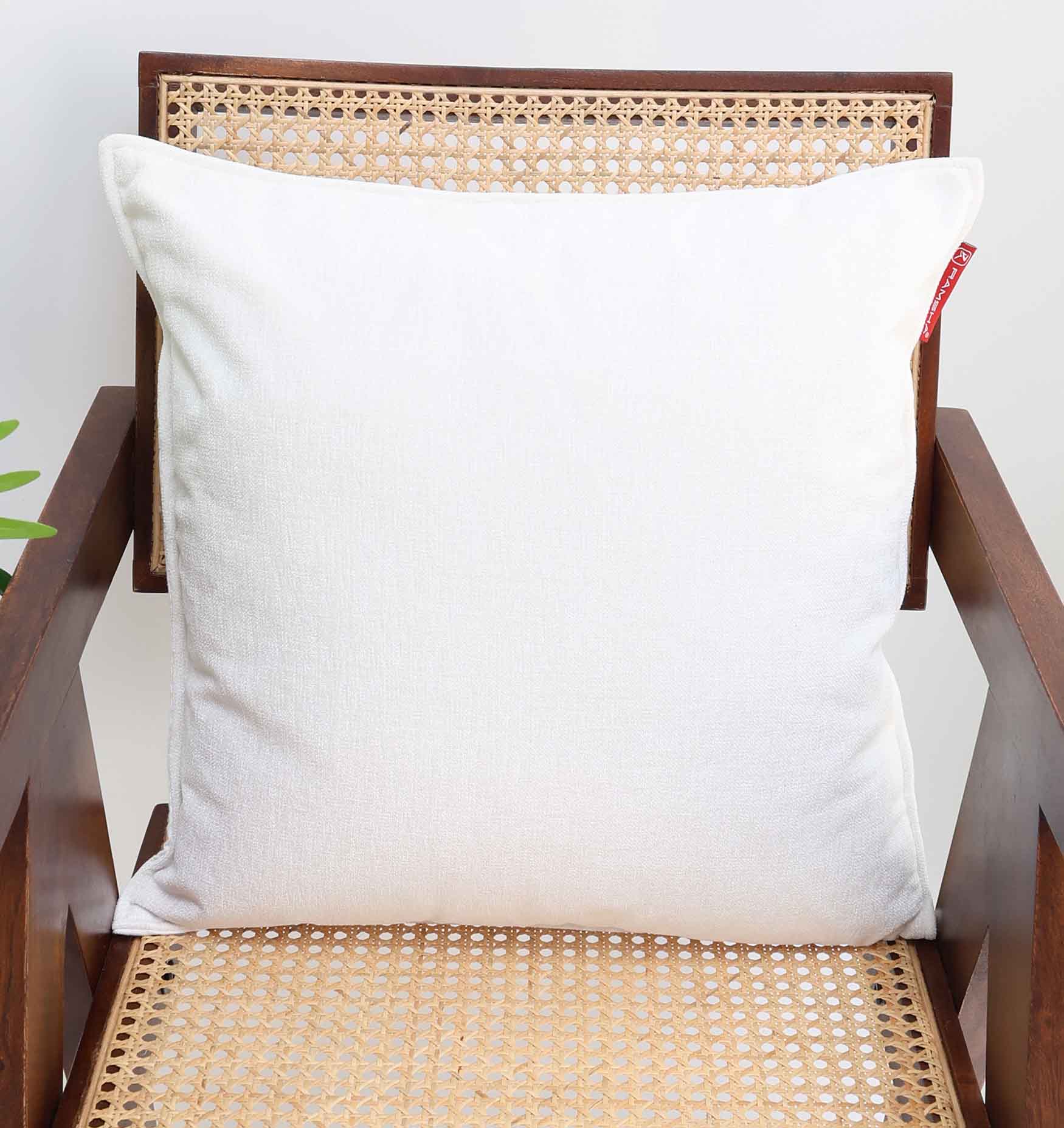 The Ramsha Home White Color Decorative Cushion For Sofa (CS 293) sits on a wooden chair with woven cane, its subtle red tag adding charm to the scene. A green plant peeks in from the left, enhancing this inviting home décor setting.