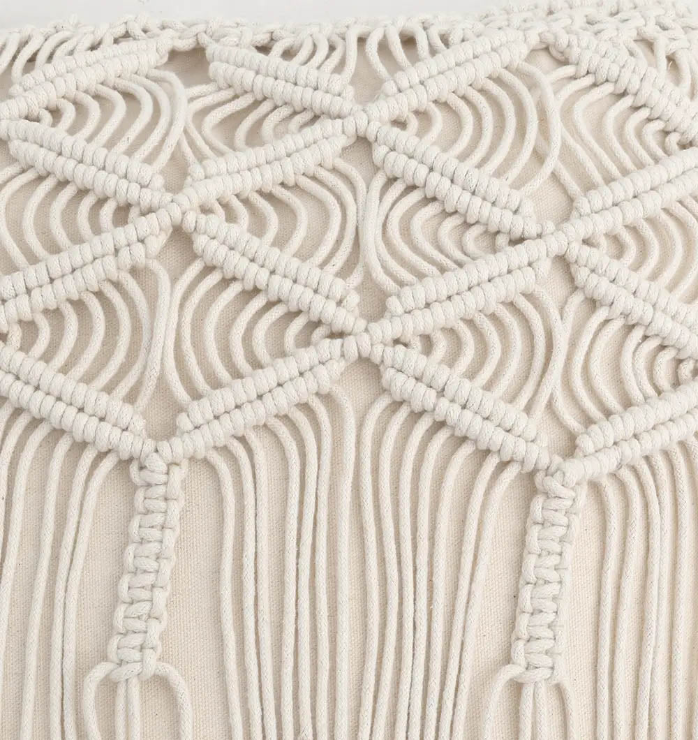 A close-up of the Natural White Macrame Sofa Cushion (CS 309) by Ramsha Home shows its cream macramé diamond and loop patterns with fringe, crafted from thick cotton cord, set against a matching fabric—luxurious UAE home furnishing.