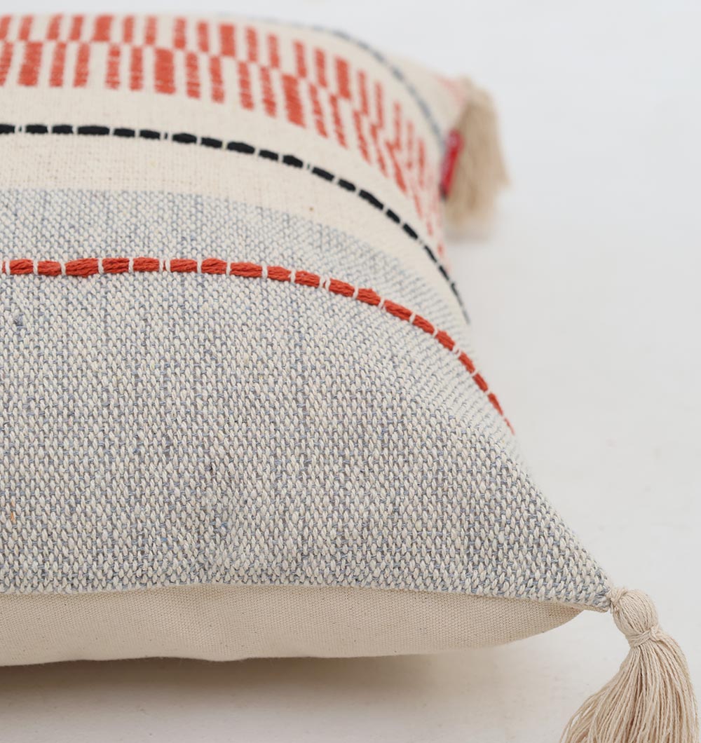 The Multi Color Home Decor Cushion - CS 314 B by Ramsha Home features red, black, and blue stitched patterns on cream cotton with decorative tassels on each corner—a stylish accent from a luxury UAE brand.