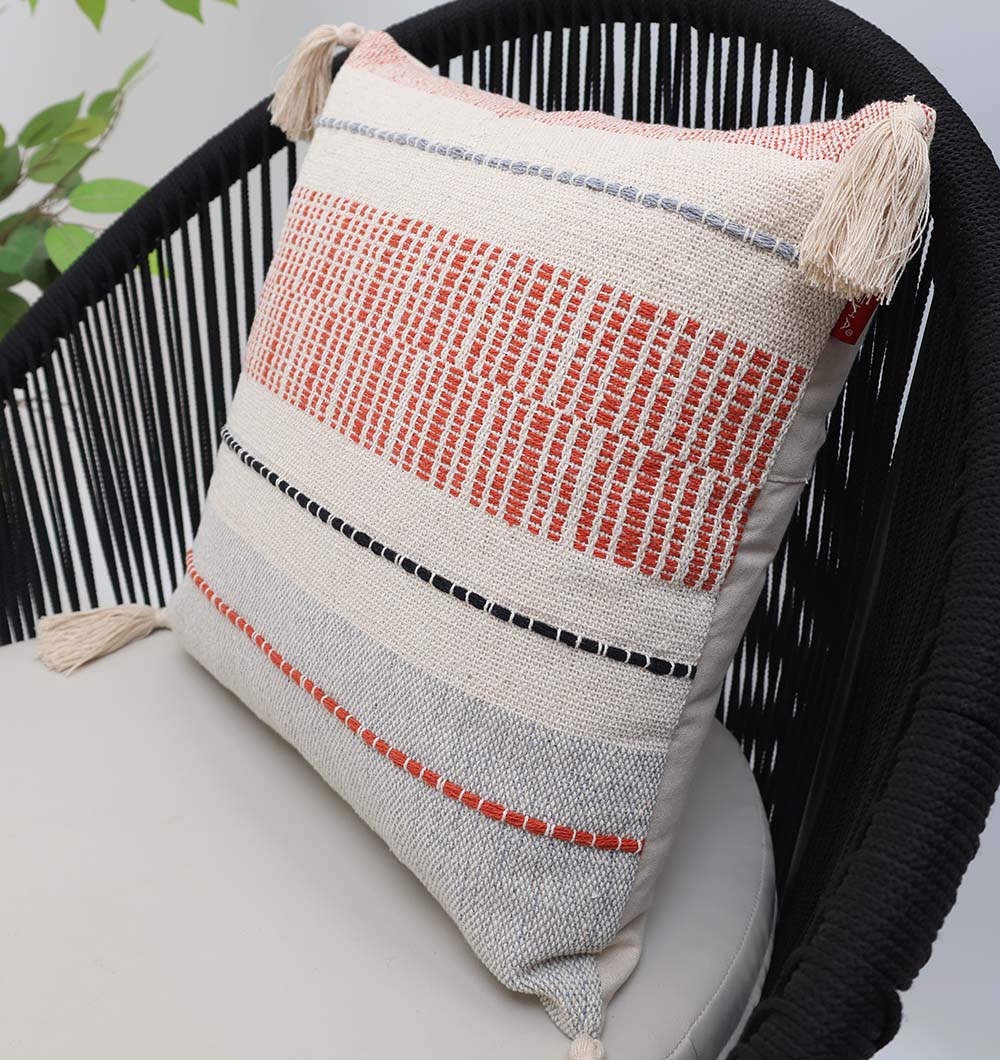 The Multi Color Home Decor Cushion - CS 314 B by Ramsha Home, featuring red, black, and gray woven patterns and tassels on each corner, sits on a rope-detailed black chair beside a green leafy plant.