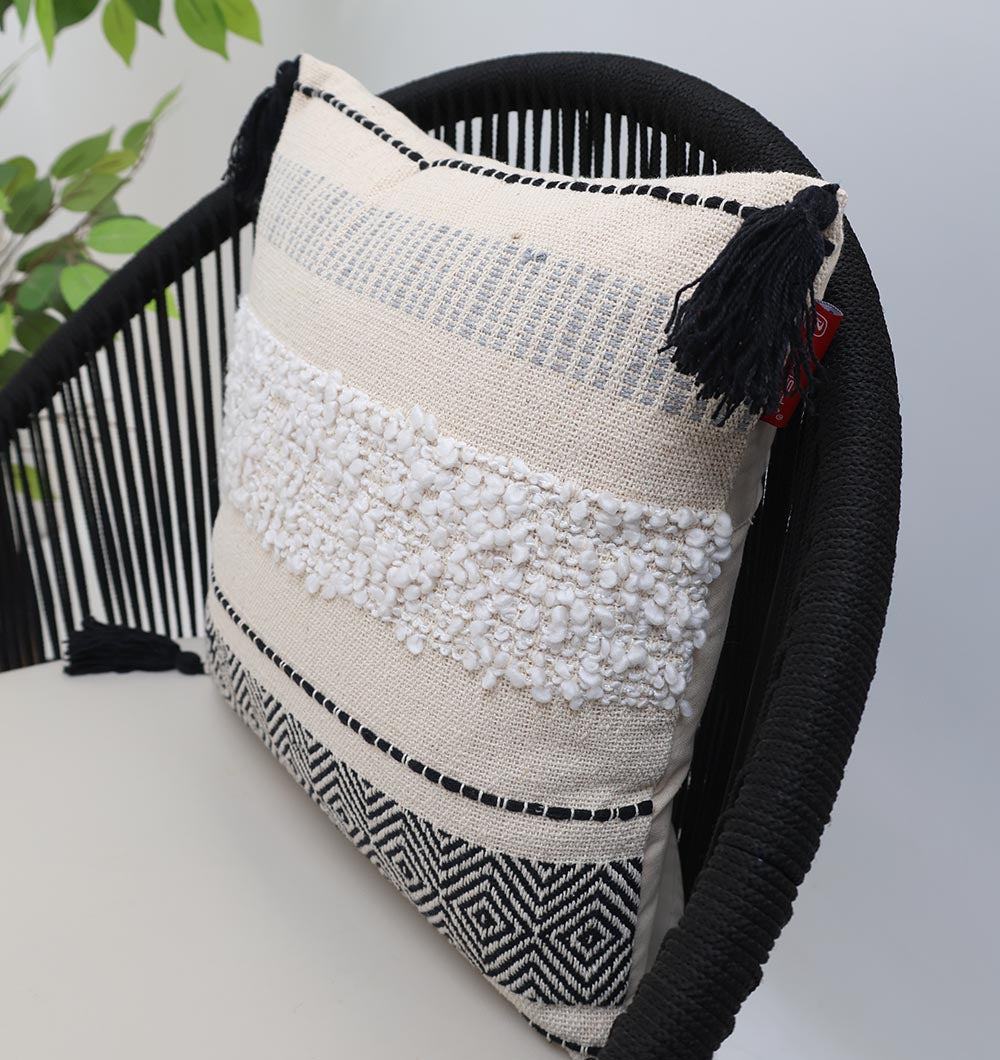 The Multi Color Home Decor Cushion - CS 314 C by Ramsha Home, a luxury UAE brand, features cream and black color blocking with tasseled corners, displayed on a modern black chair beside a green leafy plant.