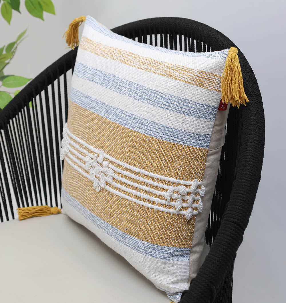 The Multi Color Home Decor Cushion - CS 315 A by Ramsha Home, featuring yellow and blue stripes, white textured patterns, and yellow corner tassels, is displayed on a black woven chair with a beige cushion. A green plant appears in the background.