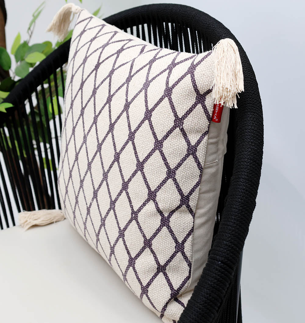 The Cream & Brown Printed Cotton Cushion 45x45 cm CS 318 by Ramsha Home features a black geometric diamond pattern and beige tassels, displayed on a modern black chair with a green plant partly visible in the background.