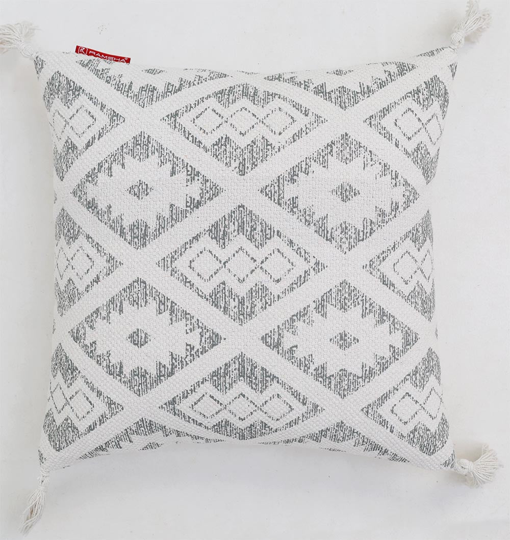 The Ramsha Home White and Grey Printed Cotton Cushion 45x45 cm (CS 319) features a geometric diamond and zigzag pattern, decorative tassels at each corner, and a plain white background.