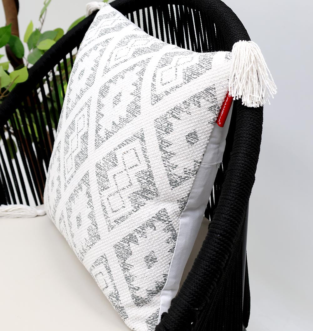 The Ramsha Home White and Grey Printed Cotton Cushion 45x45 cm (CS 319) rests on a black woven chair, featuring a geometric pattern and tassels. Green leaves are partly visible in the background.