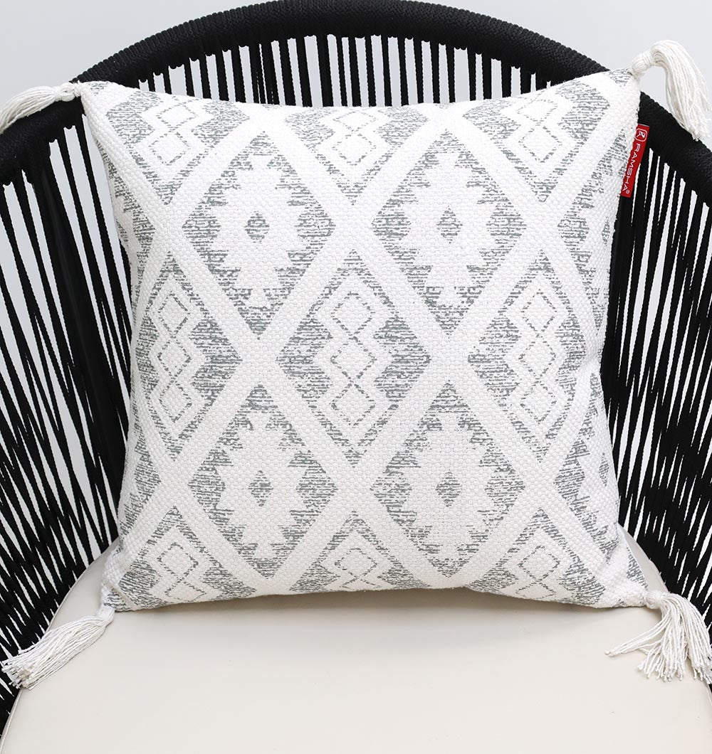 The Ramsha Home White and grey Printed Cotton Cushion 45x45 cm (CS 319) rests on a modern black wire chair with a cream seat, its geometric diamond pattern and corner tassels adding style to the decor.