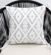 The Ramsha Home White and grey Printed Cotton Cushion 45x45 cm (CS 319) rests on a modern black wire chair with a cream seat, its geometric diamond pattern and corner tassels adding style to the decor.