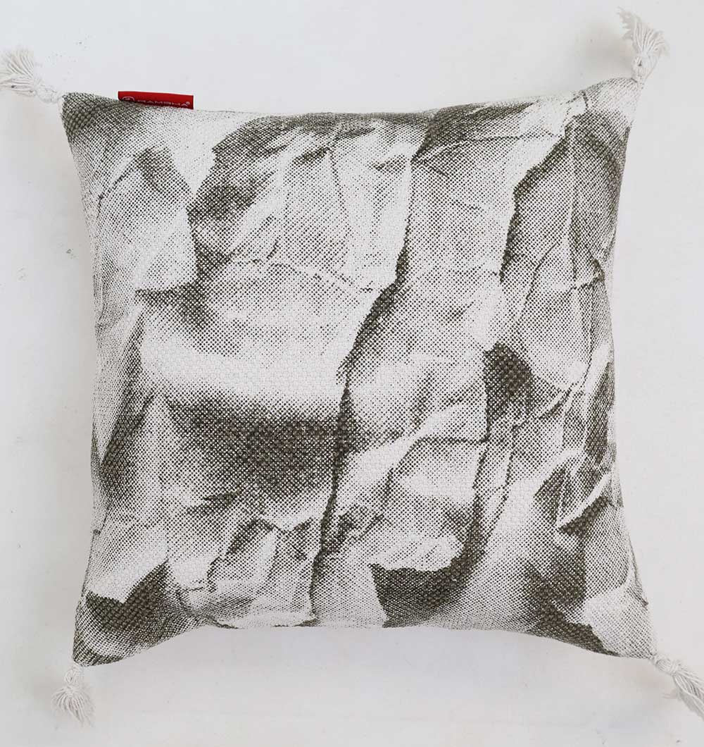 The Ramsha Home White and Grey Printed Cotton Cushion 45x45 cm CS 320 features a premium cotton build, textured black-and-white crumpled paper print, corner tassels, and a red tag on the upper left edge.