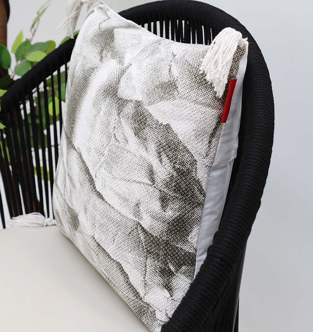 A black woven chair with a beige cushion displays the Ramsha Home White and Grey Printed Cotton Cushion 45x45 cm CS 320, featuring white tassels and a red tag—a stylish accent from this luxury UAE home furnishings brand amid green leaves.