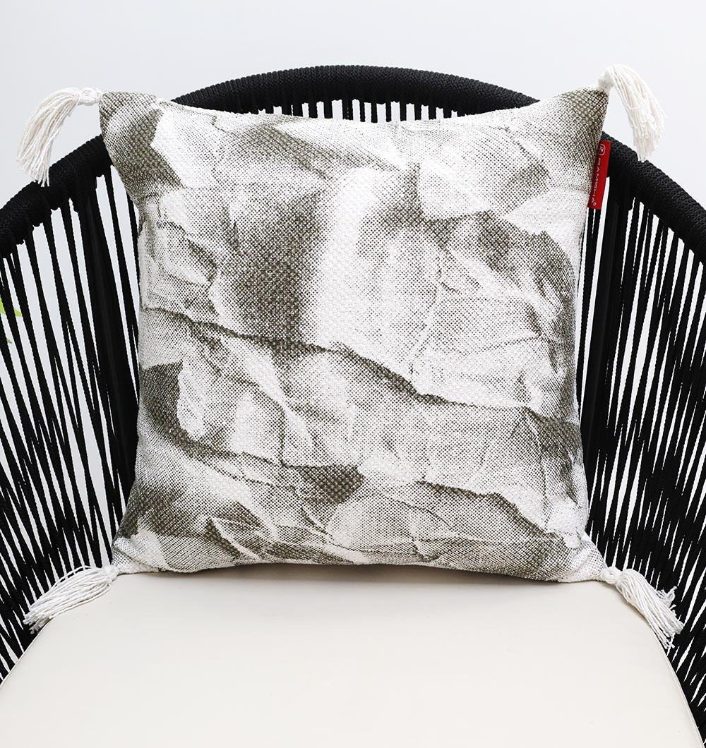 The Ramsha Home White and Grey Printed Cotton Cushion 45x45 cm CS 320 features a crumpled paper pattern, white tassels, and adds elegance to any space. Shown on a black chair with woven sides and a cream seat.