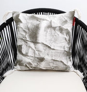 The Ramsha Home White and Grey Printed Cotton Cushion 45x45 cm CS 320 features a crumpled paper pattern, white tassels, and adds elegance to any space. Shown on a black chair with woven sides and a cream seat.