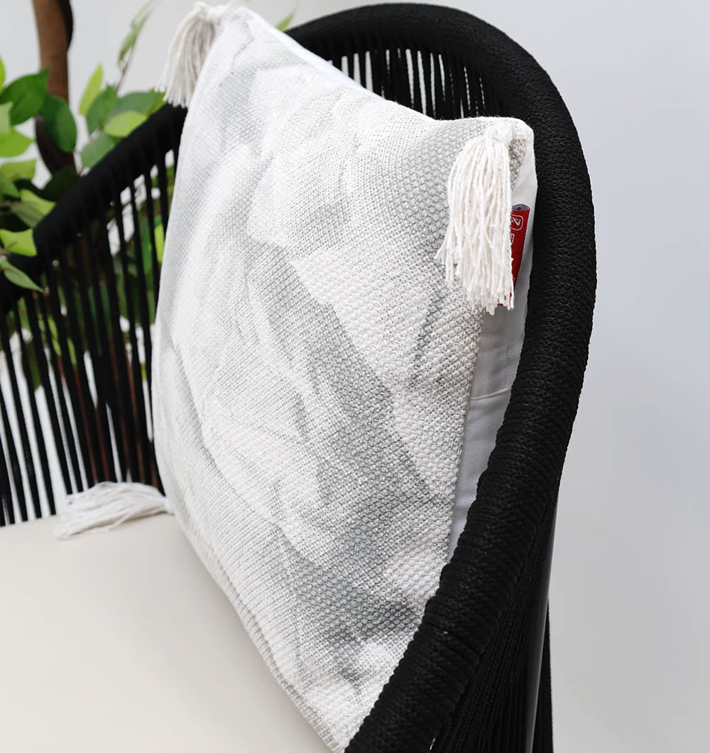 The Ramsha Home White and Grey Printed Cotton Cushion 45x45 cm (CS 321) adds style to a black woven chair, with green leaves from a nearby plant in the background.