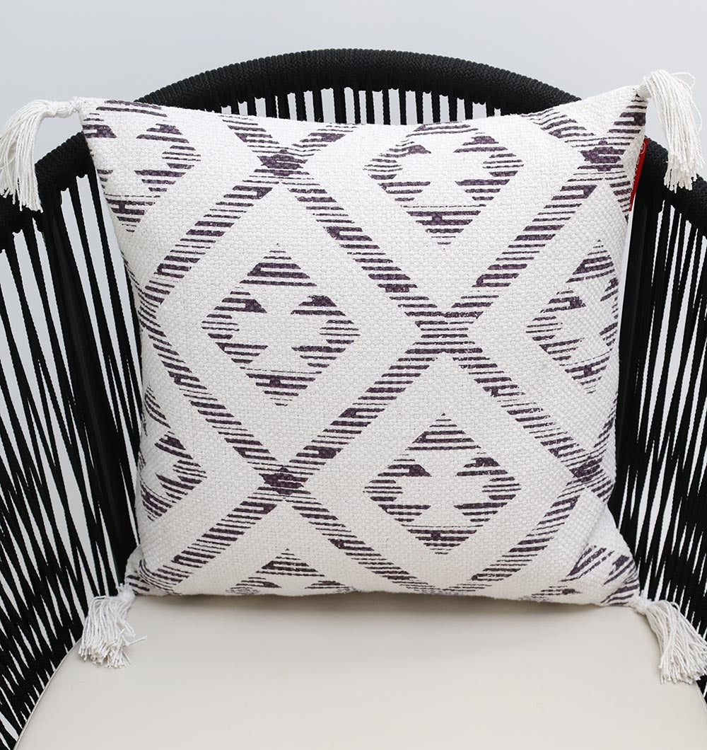 The Ramsha Home White and Grey Printed Cotton Cushion 45x45 cm CS 322 adds elegance to a black chair with vertical slats, featuring geometric diamond patterns and decorative tassels on the corners.