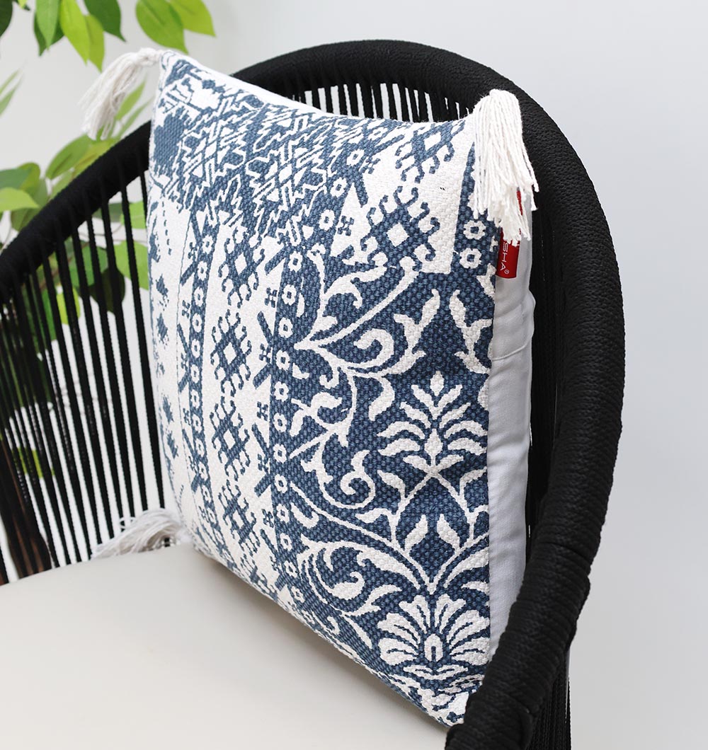 The White and Blue Printed Cotton Cushion 45x45 cm CS 323 by Ramsha Home adds elegance to a black woven chair with a beige seat, while a green plant enhances the serene ambiance. Perfect for chic interiors from this luxury UAE brand.