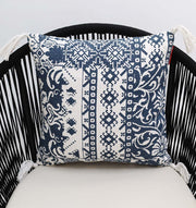 The Ramsha Home White and Blue Printed Cotton Cushion 45x45 cm CS 323, with geometric and floral motifs plus tasseled corners, adds elegance when placed on a black vertical-slat chair.