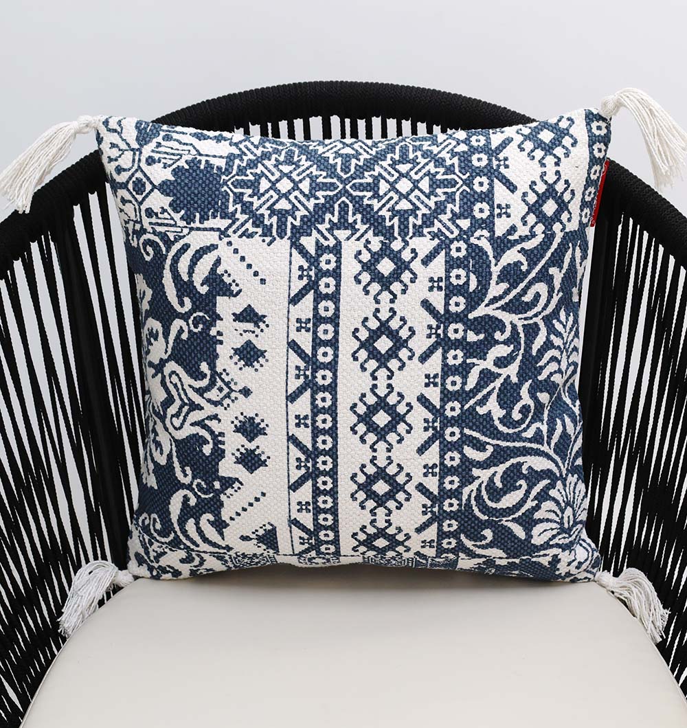The Ramsha Home White and Blue Printed Cotton Cushion 45x45 cm CS 323, with geometric and floral motifs plus tasseled corners, adds elegance when placed on a black vertical-slat chair.