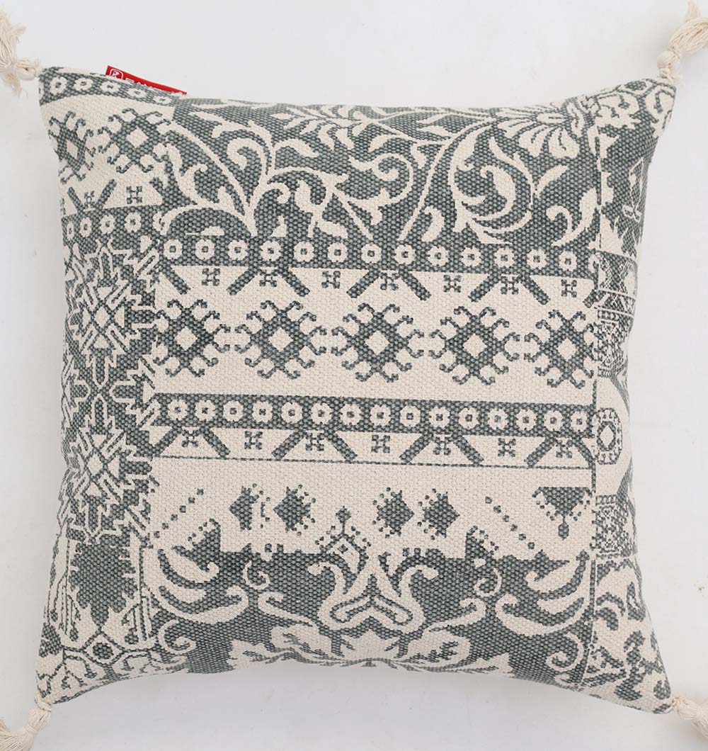 The Ramsha Home White and Grey Printed Cotton Cushion 45x45 cm (CS 324) features geometric and floral patterns, corner tassels, and a red tag for a stylish touch to your décor.